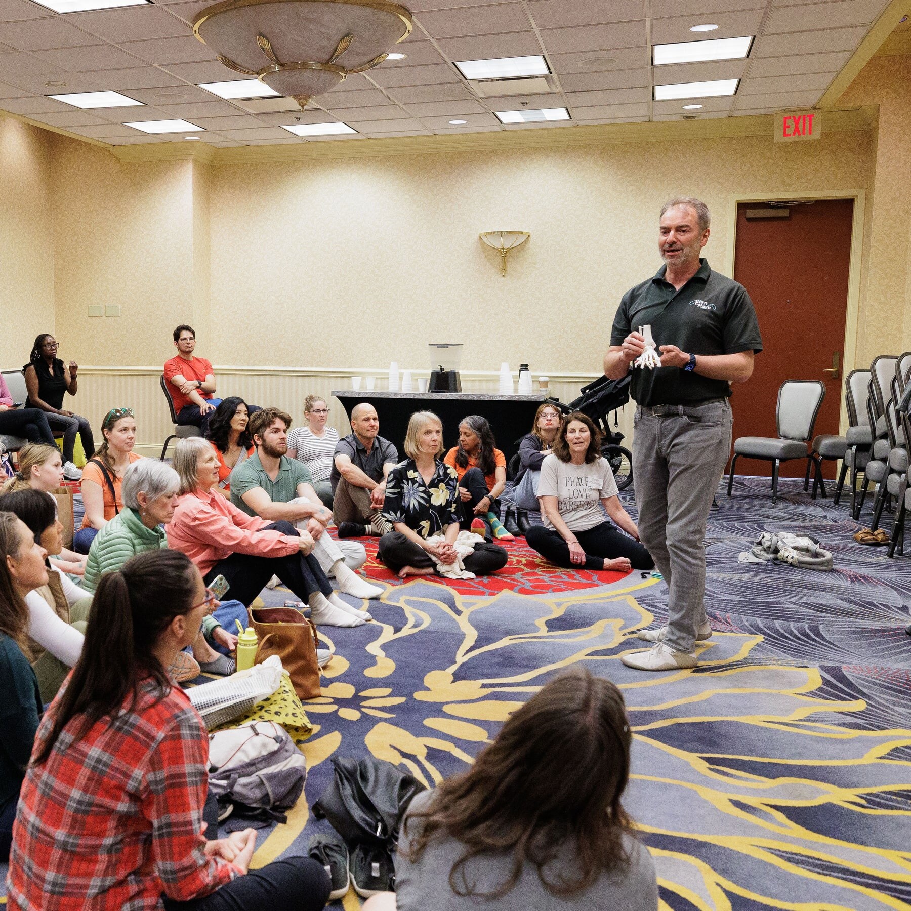 A presenter speaking to workshop participants seated on the floor, photo by Barefoot Shoe Expo