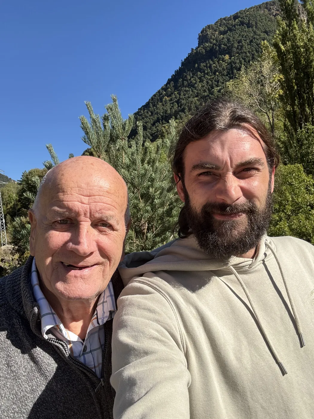 Dan and his grandad smiling in the sunshine with a pine covered mountain behind