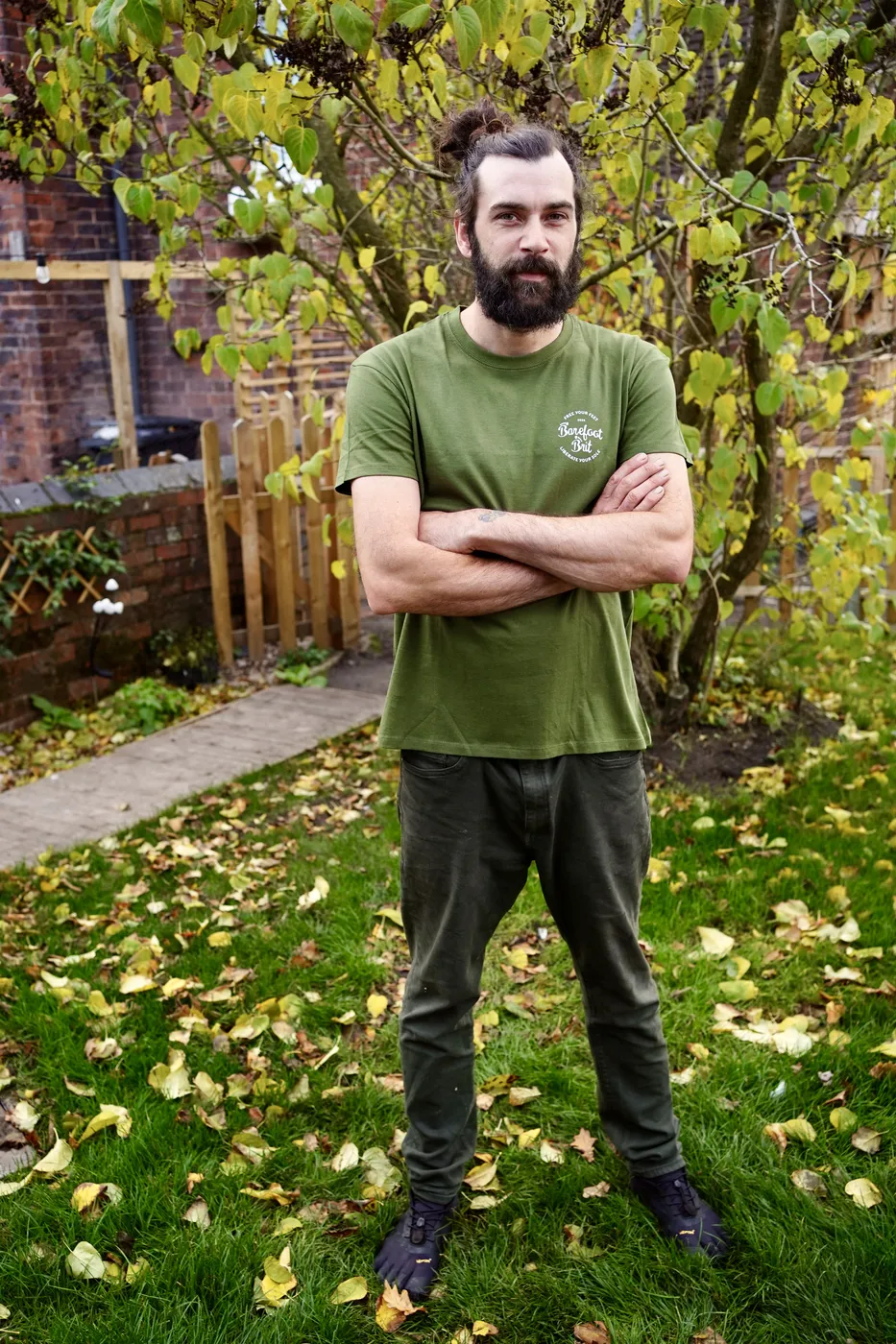 Dan standing in the garden in a green Barefoot Brit t-shirt and toe shoes, arms folded, autumn leaves on the grass